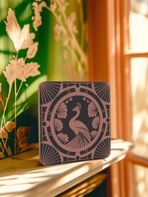 Square ceramic coaster featuring a detailed Art Deco peacock design in copper tones, surrounded by shell motifs and geometric fan patterns on a dark charcoal background, with protective cork base.